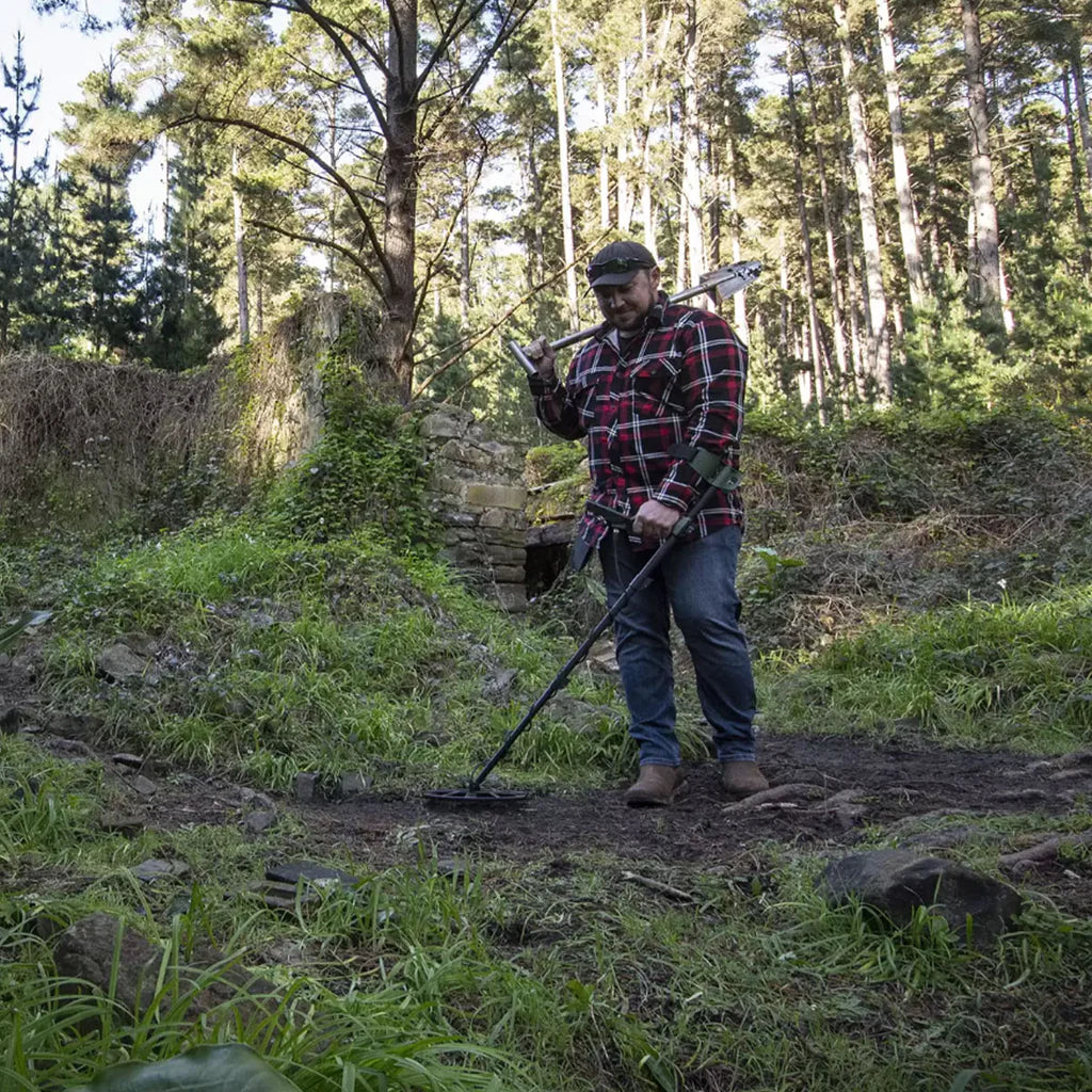 Una persona con ropa de aventura y mochila, sosteniendo un detector de metales X-TERRA PRO plegado y compacto, listo para ser transportado a un sitio de búsqueda remoto. El fondo muestra un paisaje natural con árboles y un sendero, enfatizando su diseño ergonómico y portátil.